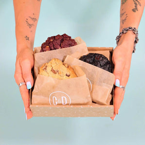 Hands holding a box with 3 x New York Cookies, The Classics including chocolate chip, double chocolate, and red velvet flavors from Crumbs & Doilies.